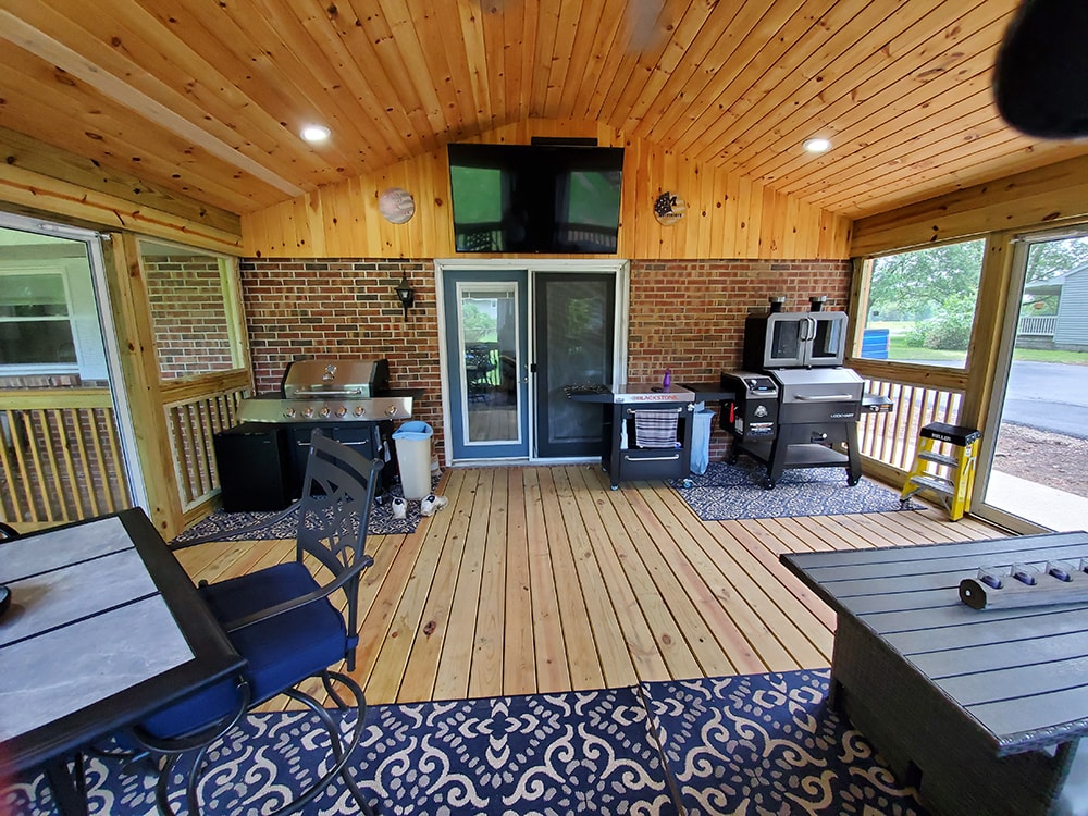 Deck addition Findlay — after: interior outdoor kitchen wall showing mounted TV, gas grill, Blackstone griddle, smoker, brick wall, and knotty pine vaulted ceiling, Findlay, Ohio