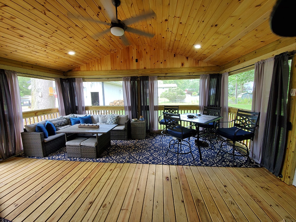 Deck addition Findlay — after: interior of screened room with knotty pine T&G vaulted ceiling, ceiling fan, recessed lights, wicker sectional sofa, dining set, and blue patterned rug, Findlay, Ohio