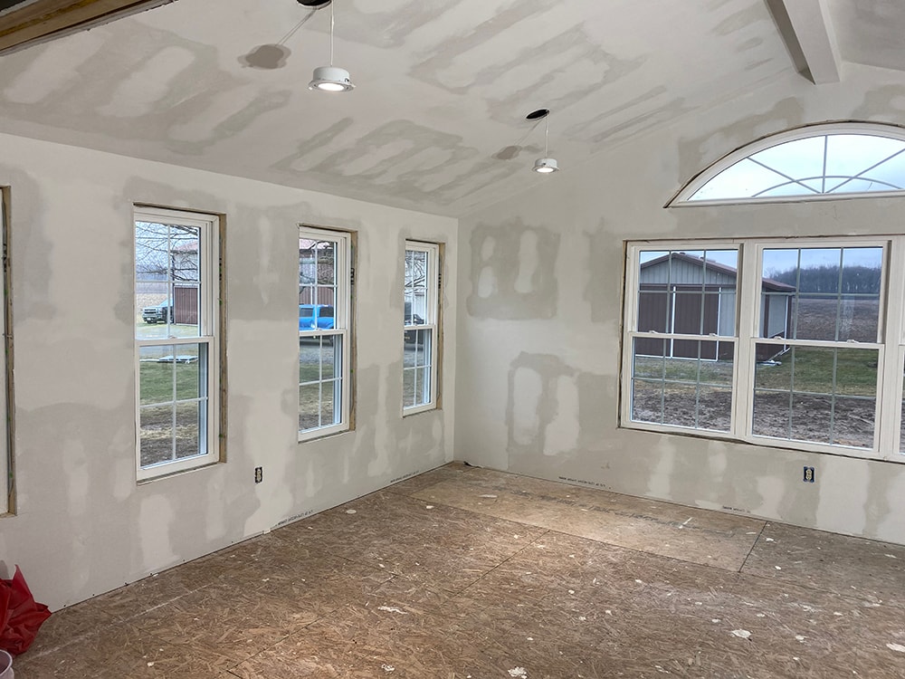 Room addition Fostoria — interior drywall mudded, opposite wall angle showing three double-hung windows and vaulted ceiling with recessed lights, Fostoria, Ohio