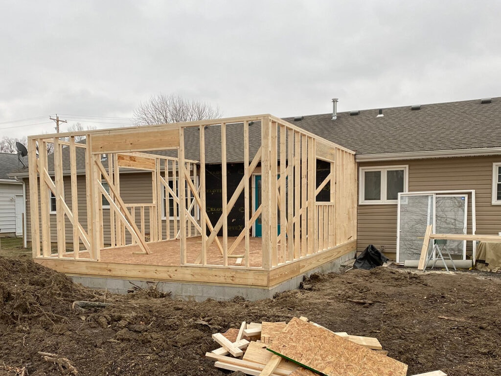 Large room addition Findlay — framing stage: full stud wall framing on new slab with OSB subfloor, teal door visible on existing home, Findlay, Ohio