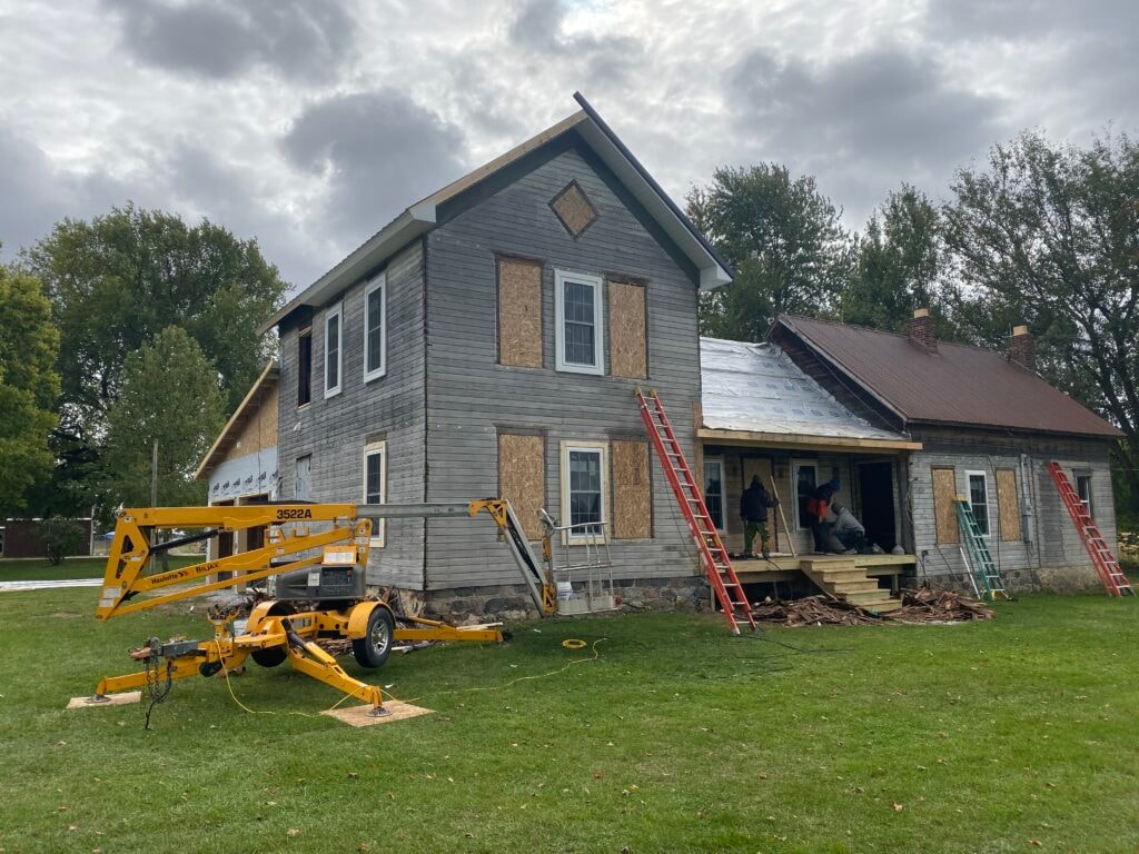Metal roof large addition Gibsonburg — during: front of home with boarded windows, new OSB sheathing, yellow lift on site, and crew working on porch, cloudy sky, Gibsonburg, Ohio