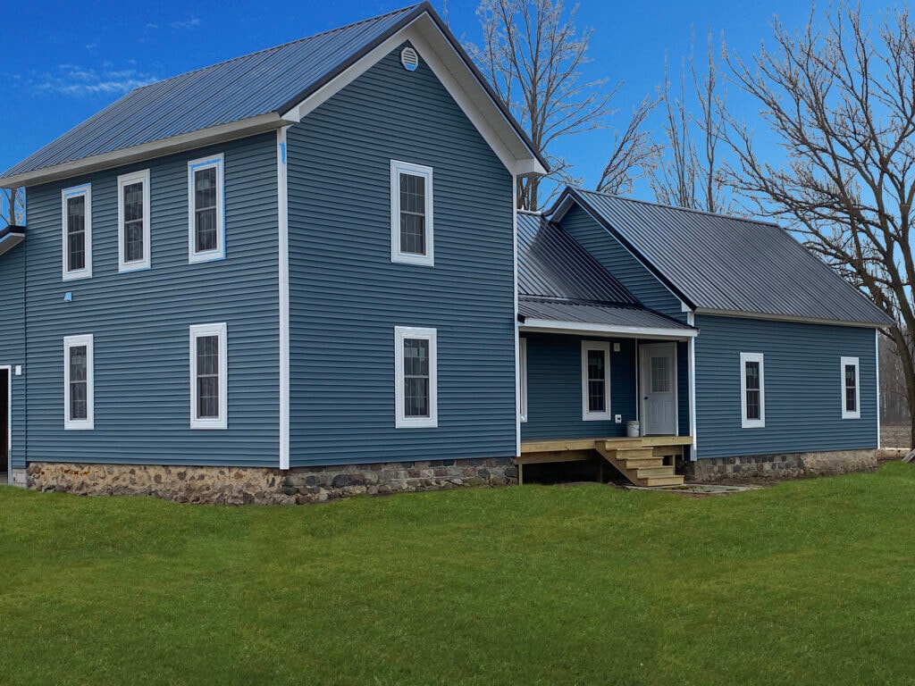 Metal roof large addition Gibsonburg — after: front of completed home with blue vinyl siding, metal roof, white trim, new entry door and steps, stone foundation, blue sky, Gibsonburg, Ohio