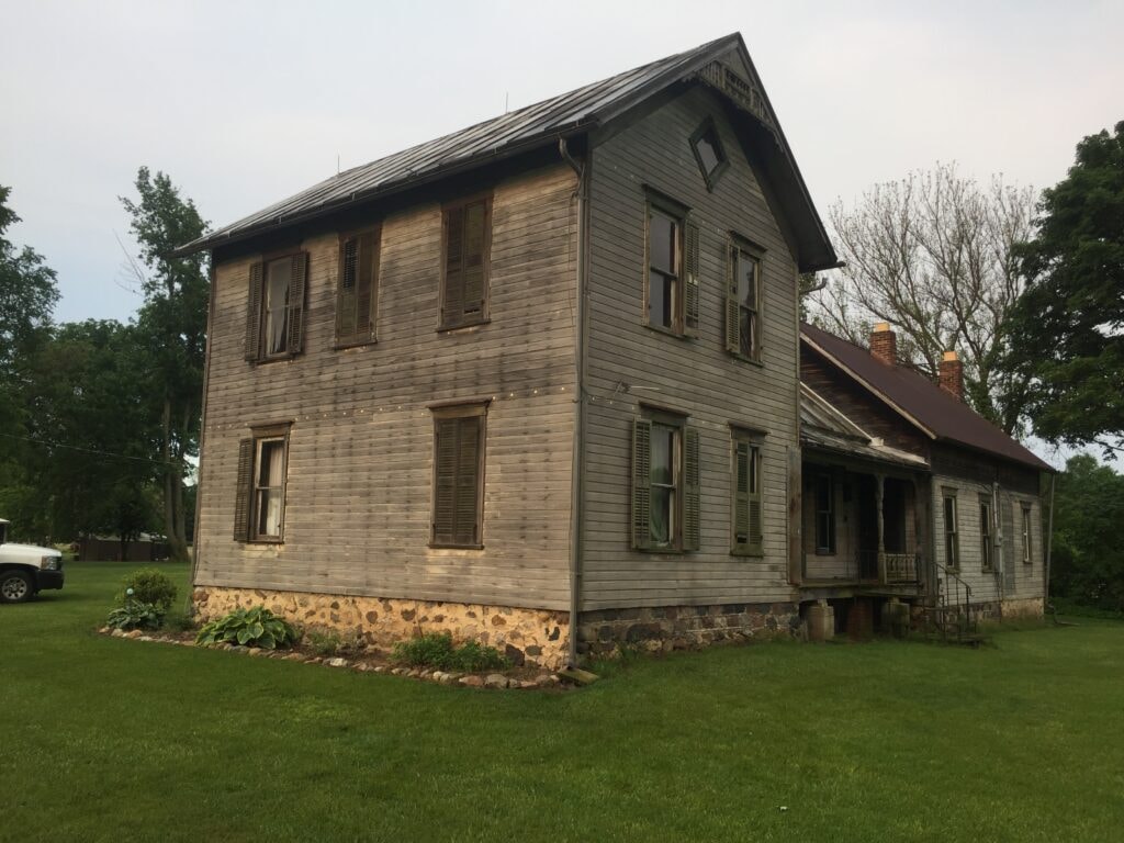 Metal roof large addition Gibsonburg — before: side view of original farmhouse with weathered wood siding, shutters, stone foundation, and attached wing, summer evening, Gibsonburg, Ohio