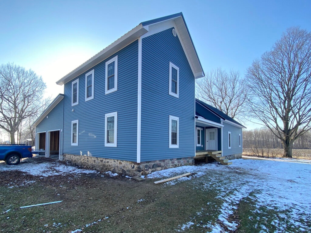 Metal roof large addition Gibsonburg — after: side of completed home with blue vinyl siding, metal roof, white trim, new entry door and steps, stone foundation, winter light, Gibsonburg, Ohio