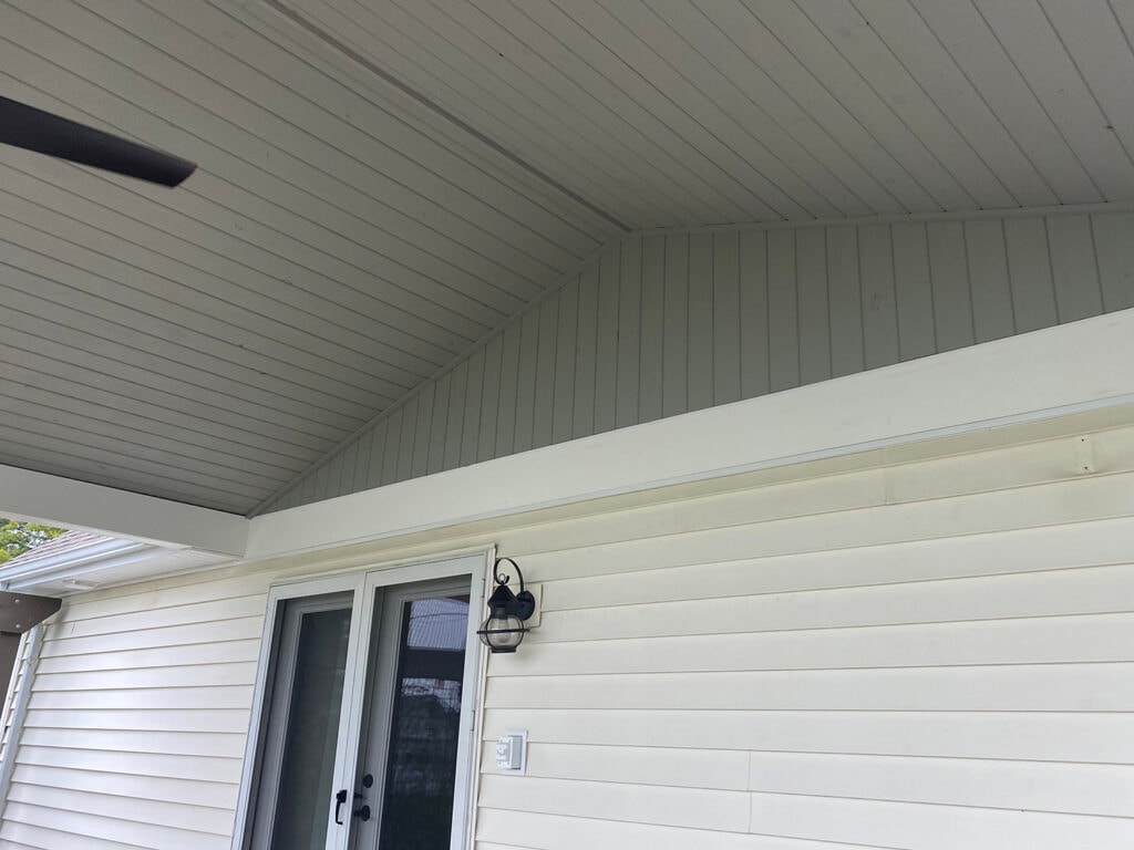 Patio addition Fostoria — after: interior close-up of tongue-and-groove beadboard ceiling, board-and-batten gable end wall, white fascia, and black lantern wall sconce beside patio door, Fostoria, Ohio