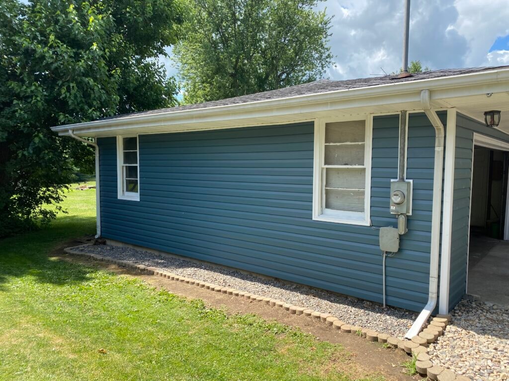 Siding job Findlay — after: side of home showing completed blue vinyl siding, white fascia, white gutters, two double-hung windows, and gravel border, Findlay, Ohio