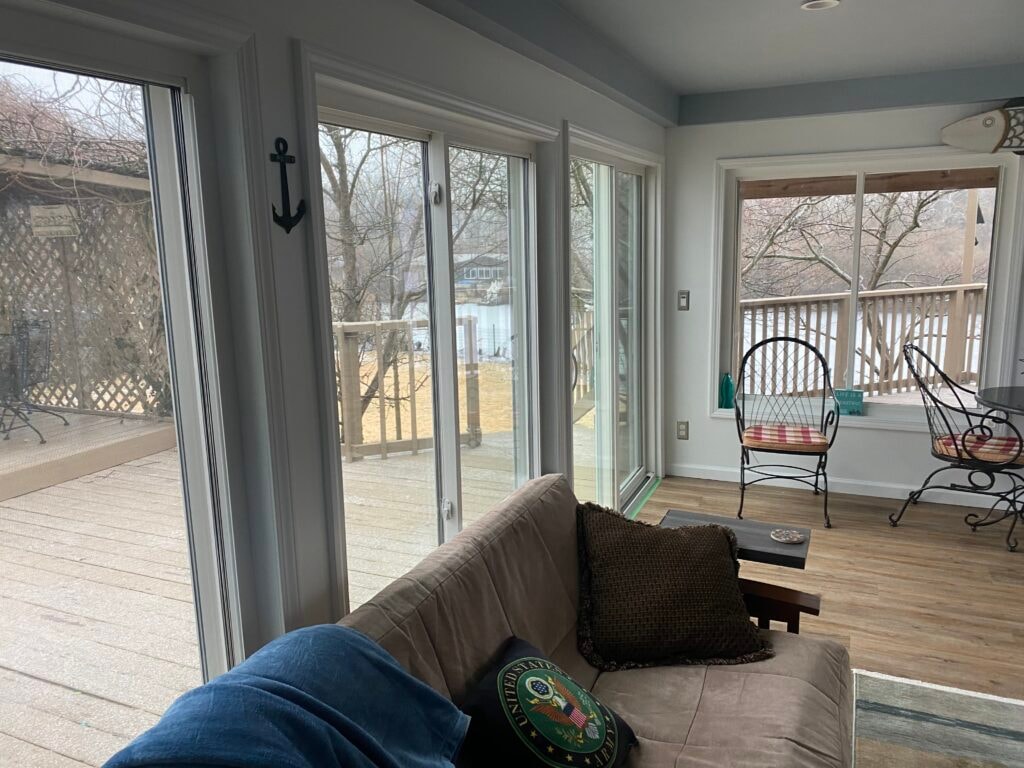 Sunroom transformation Fostoria — after: interior wide view of four-season room with floor-to-ceiling sliding glass doors, picture window, sofa, and water views beyond deck, Fostoria, Ohio